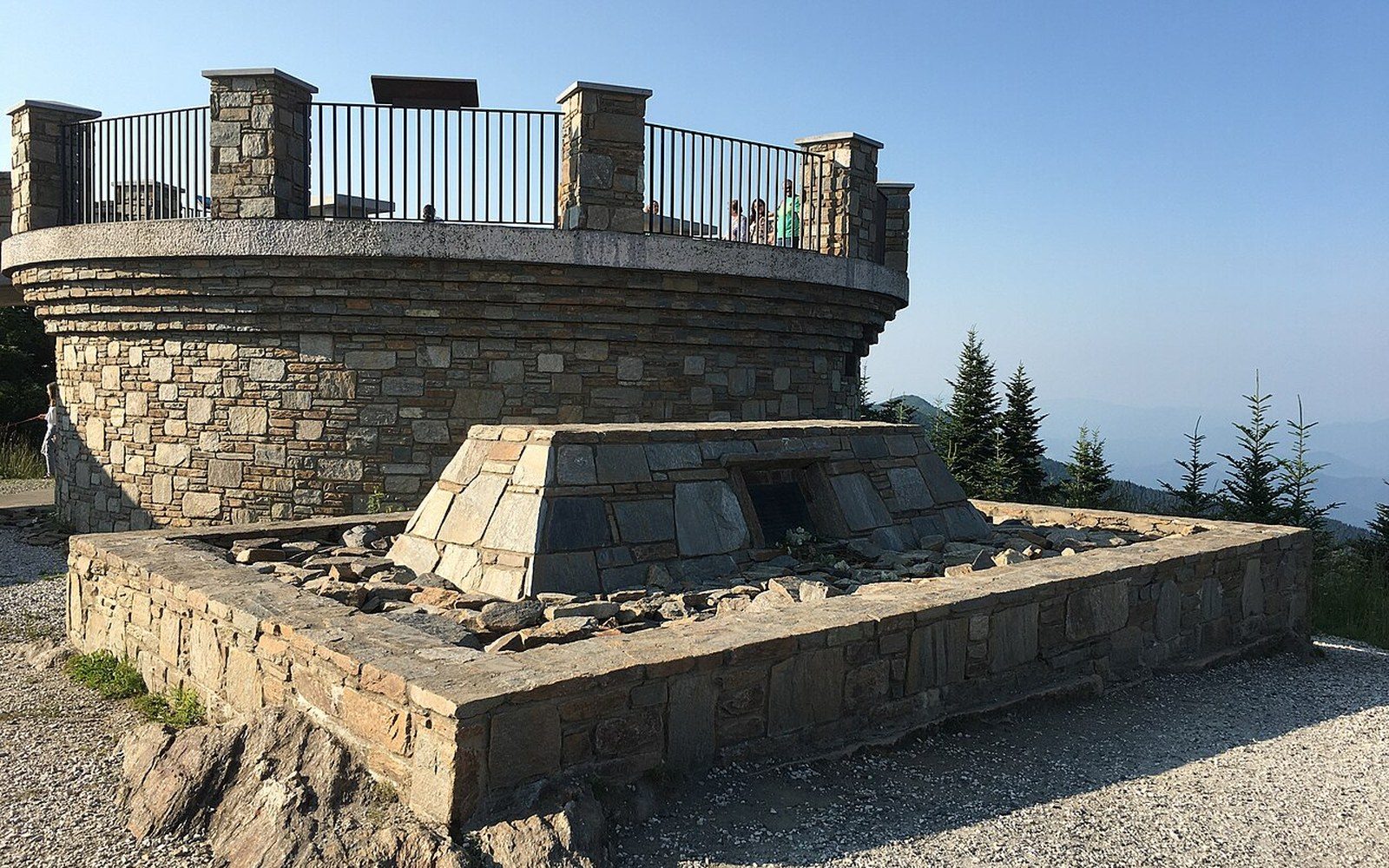 Stone observation tower and grave marker atop a mountain summit with evergreens beyond